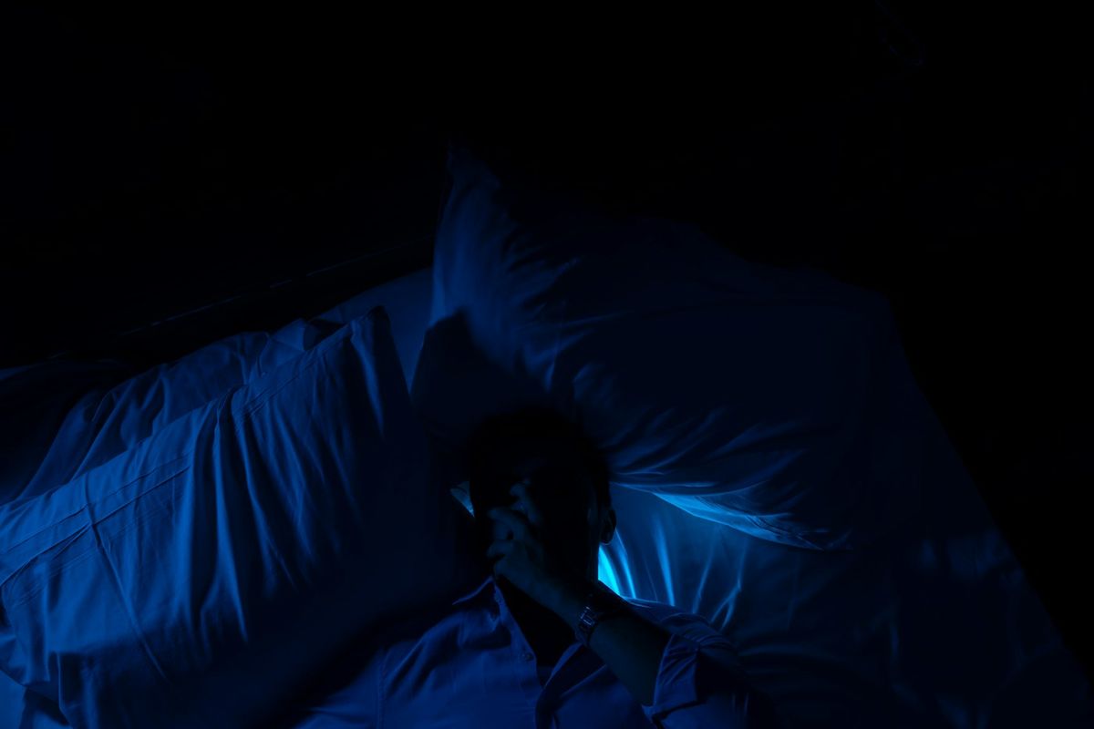 Someone lying awake in bed in a dark room, lit only by a blue glow, one hand over their face.