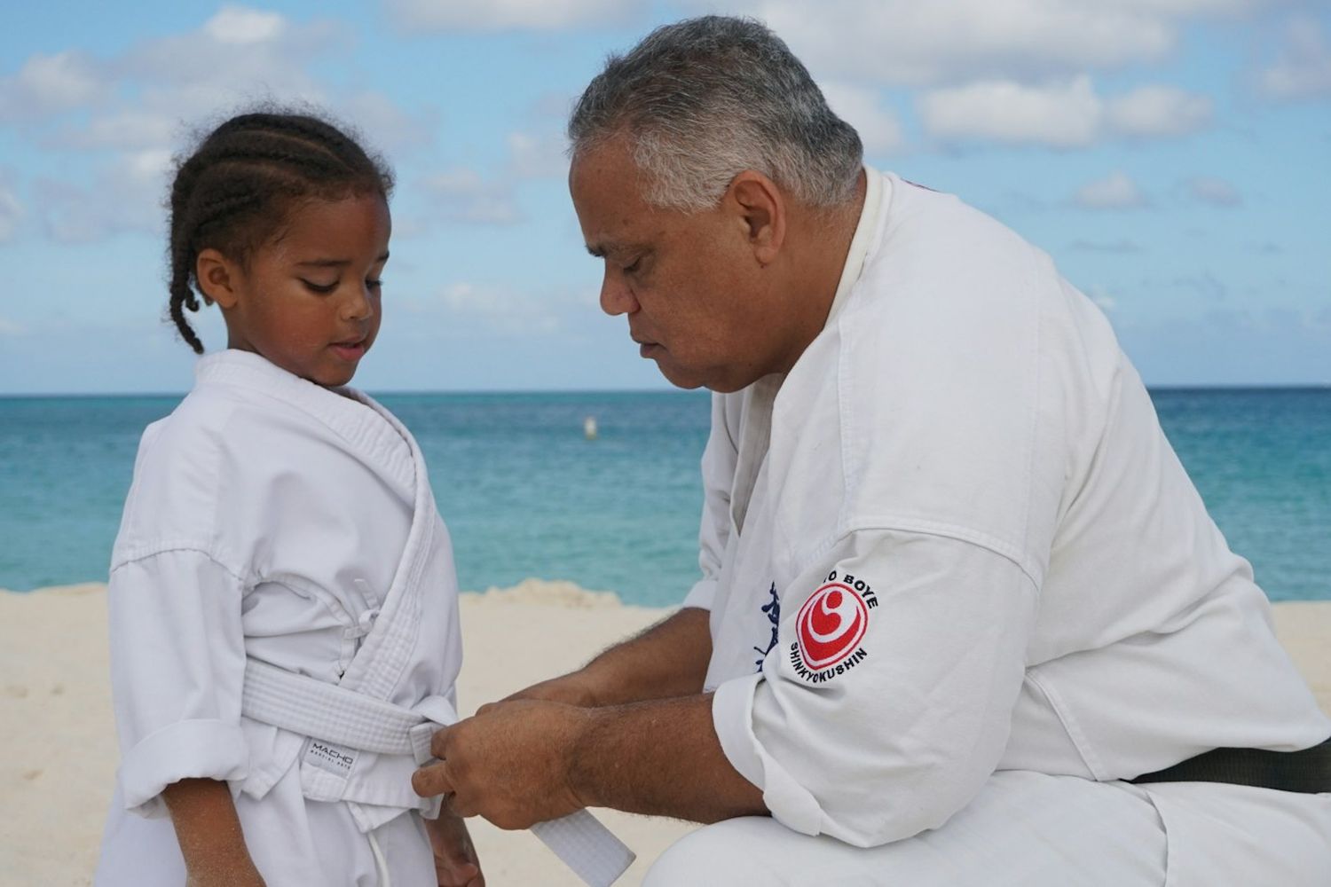 An instructor kneeling on the sand, tying a white belt on a young student.