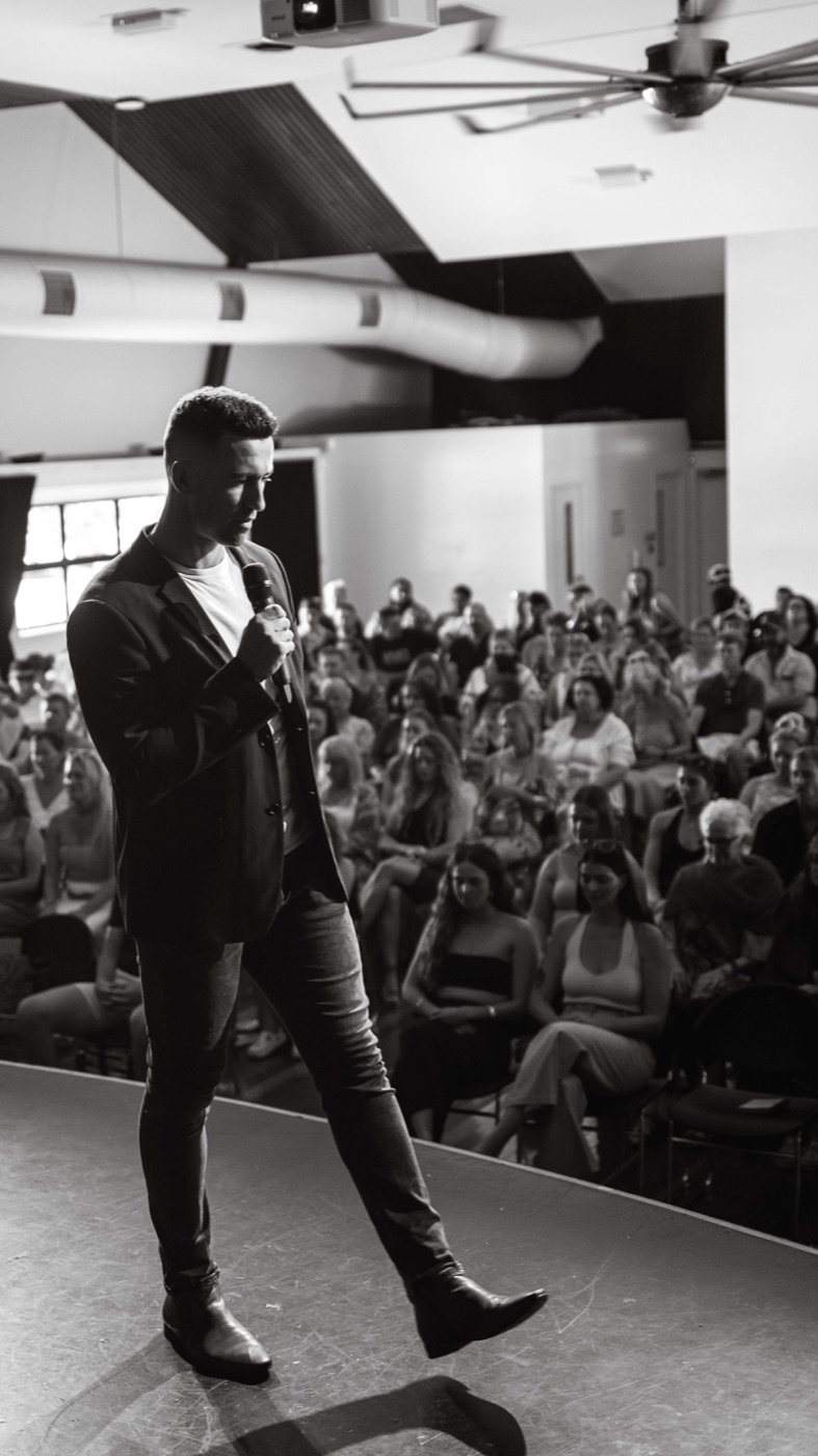 Mitch on stage with a microphone, in front of a full room.