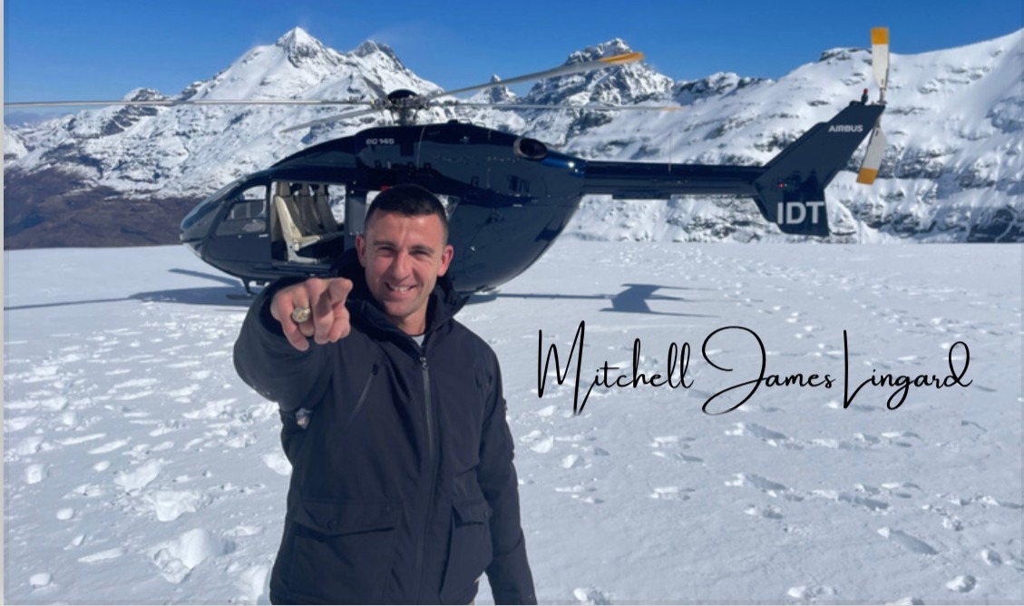 Mitch Lingard in the snow, pointing at the camera, a helicopter and mountain peaks behind him.