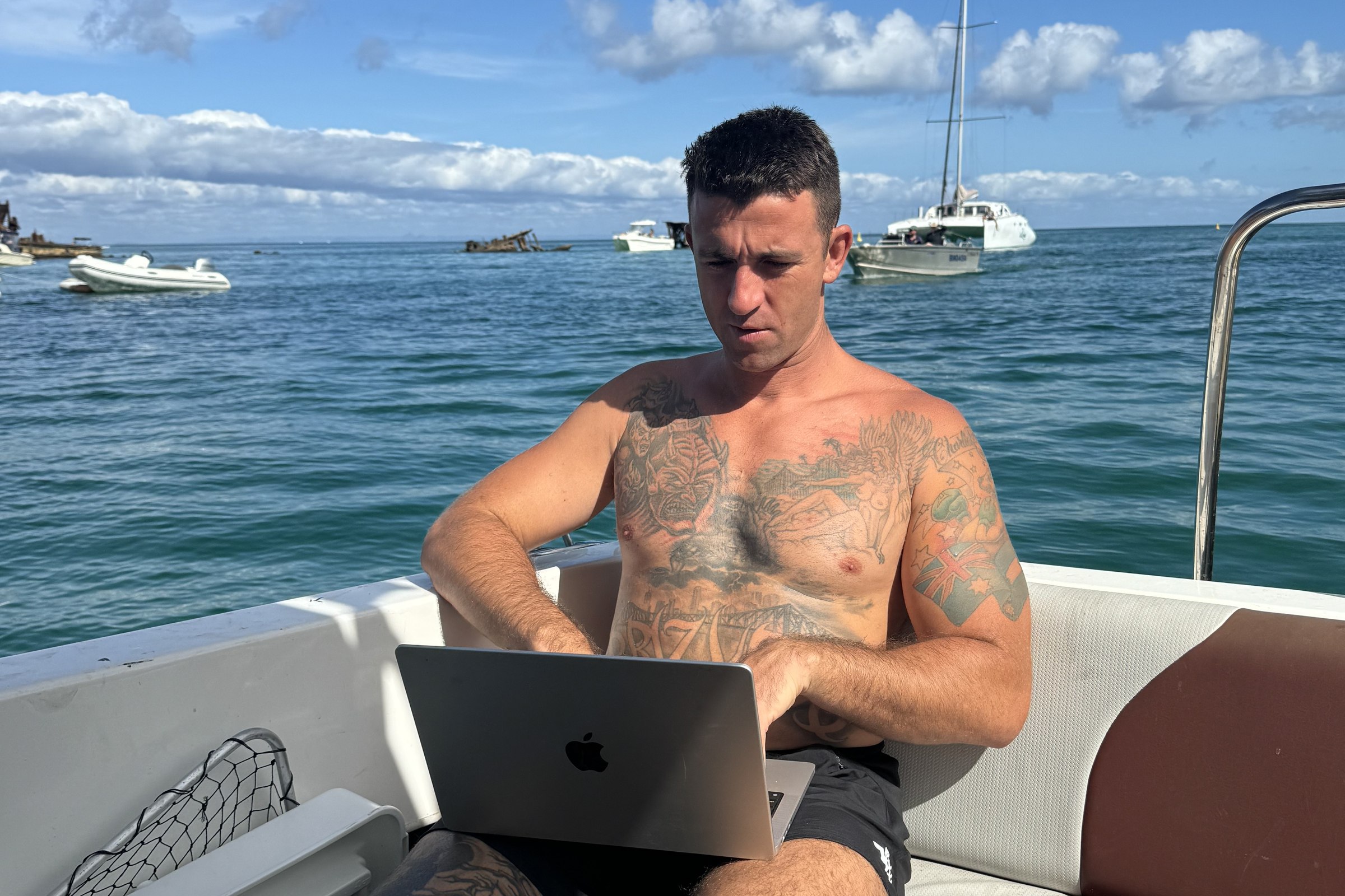 Mitch working from a boat, laptop open, the ocean and moored boats behind him.