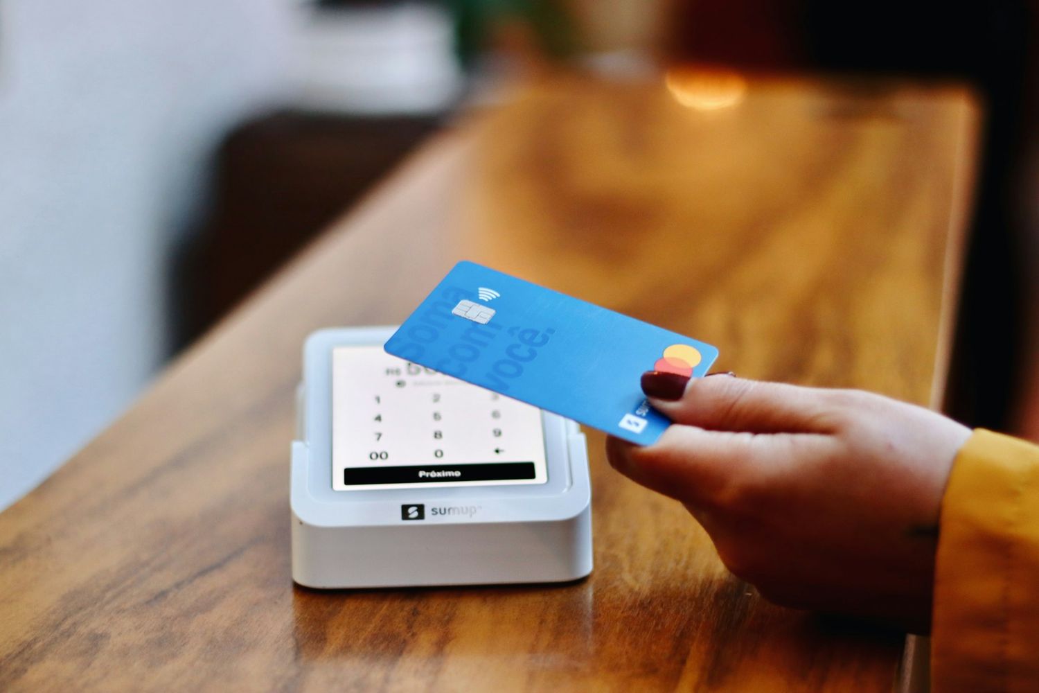 A hand tapping a blue bank card on a payment terminal on a caf&eacute; counter.